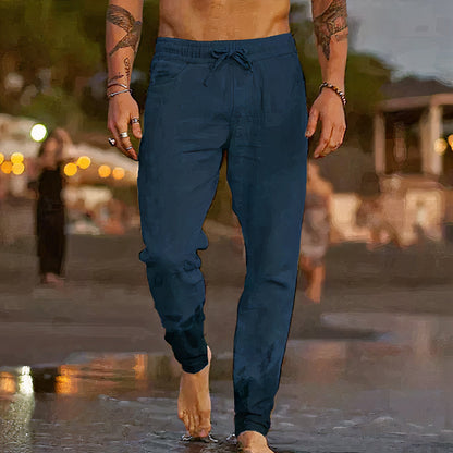 Homme torse nu portant un pantalon de jogging bleu foncé, marchant sur une plage au coucher du soleil, ambiance estivale et décontractée.