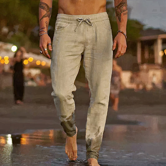 Homme torse nu portant un pantalon beige sur une plage au coucher du soleil, pieds nus dans l'eau, ambiance estivale et décontractée.
