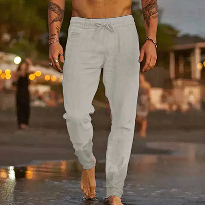 Homme torse nu marchant sur la plage au coucher du soleil, portant un pantalon de jogging gris clair, ambiance estivale et décontractée.