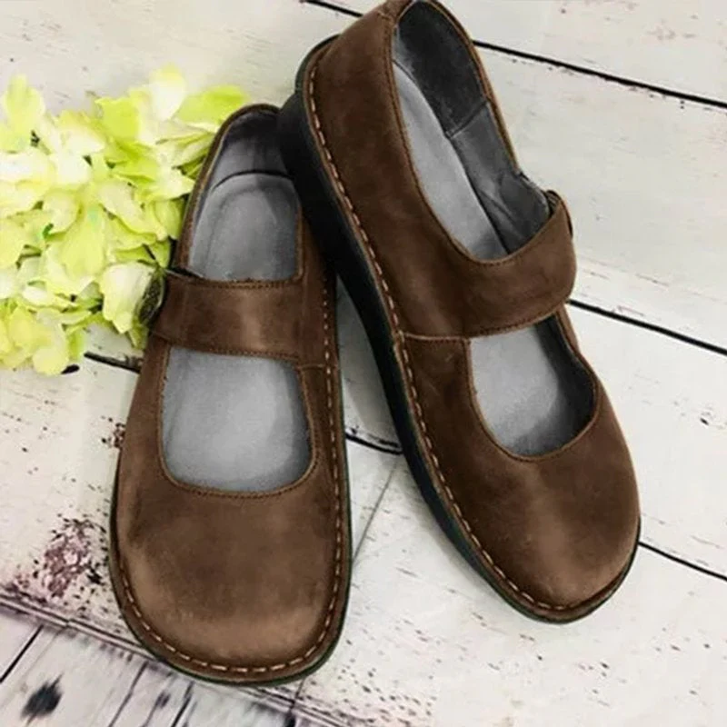 Chaussures marron en cuir style Mary Jane sur fond en bois clair, avec fleurs vertes. Confortables et élégantes, idéales pour un look décontracté.