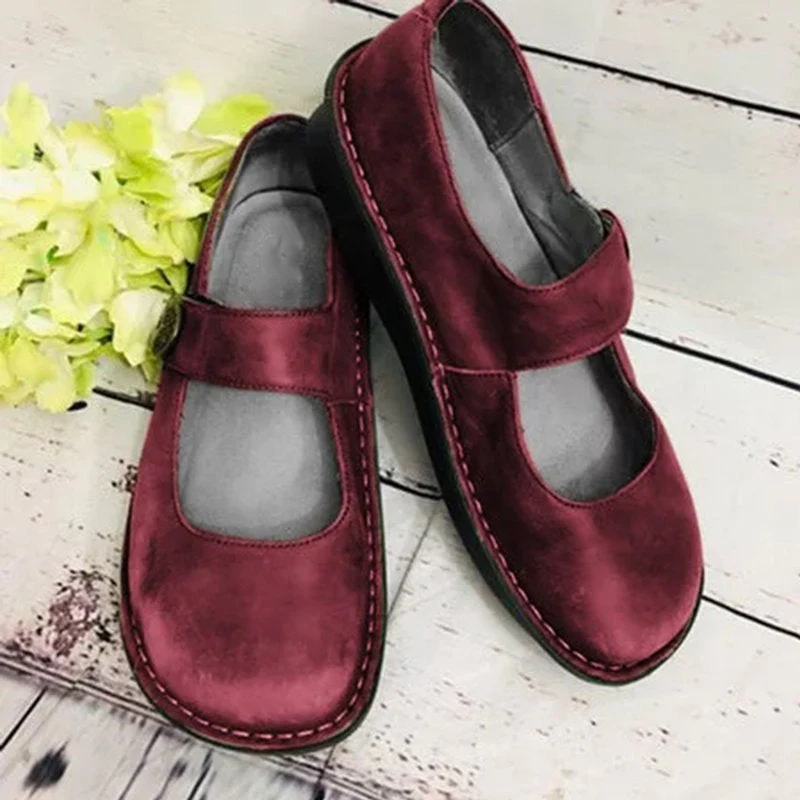 Chaussures Mary Jane bordeaux en cuir, semelle noire, posées sur un sol en bois clair avec des fleurs vertes à côté. Mode féminine élégante.