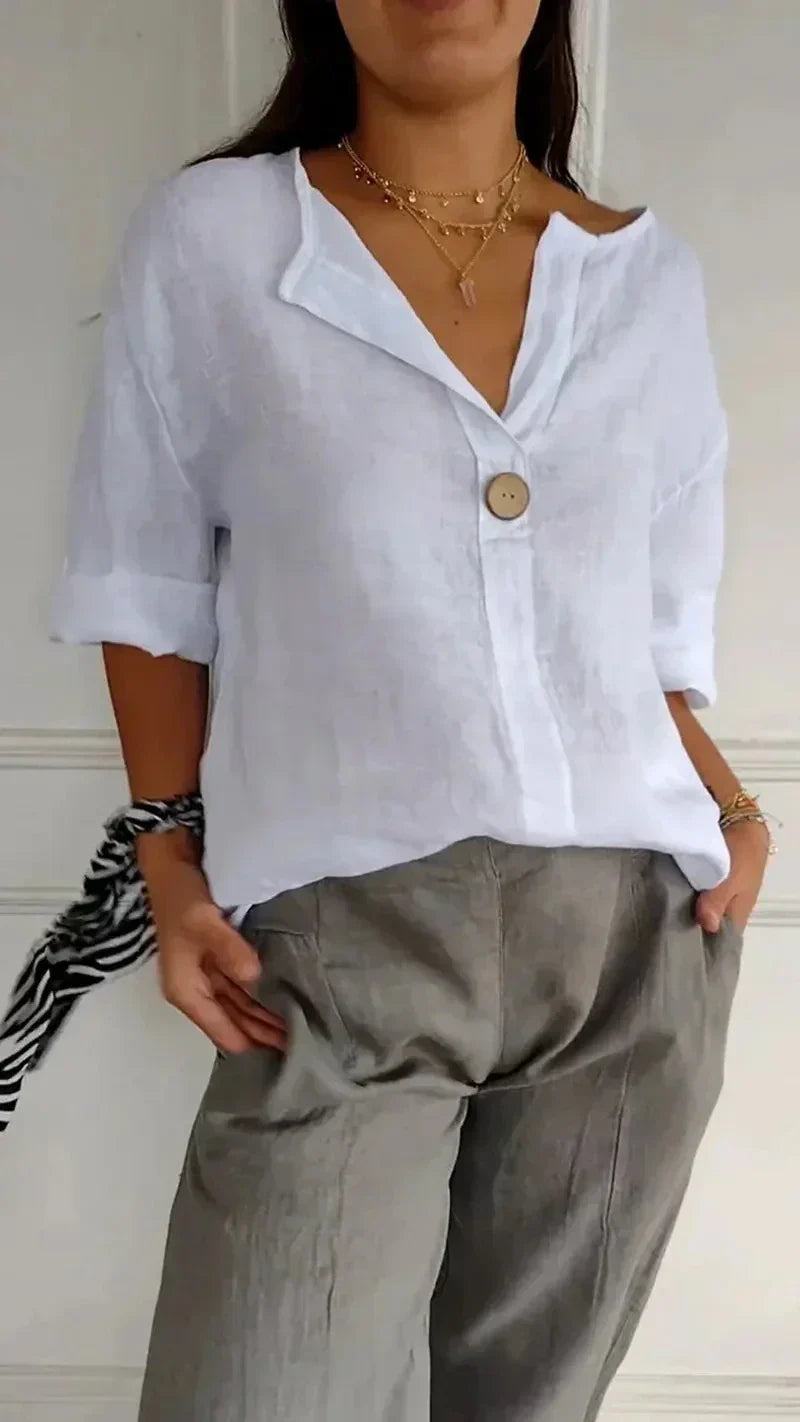 Femme portant une chemise en lin blanc avec un bouton, pantalon gris, col en V, manches retroussées, colliers dorés, style décontracté chic.
