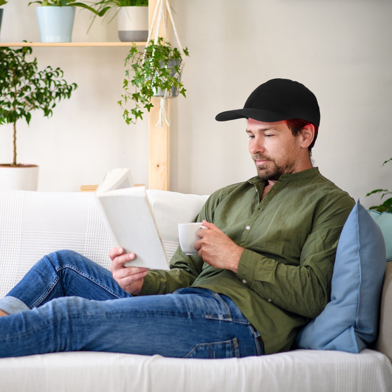 Mann liest Buch auf Sofa, trägt schwarze Kappe, grünes Hemd, blaue Jeans, hält weiße Tasse.