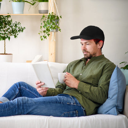 Mann liest Buch auf Sofa, trägt schwarze Kappe, grünes Hemd, blaue Jeans, hält weiße Tasse.
