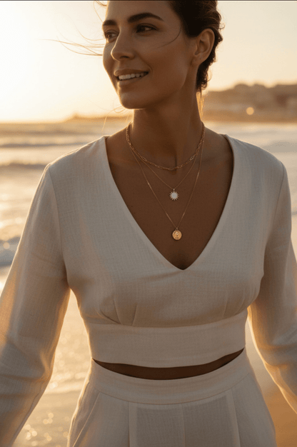 Zelfverzekerde vrouw op mediterraan strand bij zonsondergang met drie gelaagde Monerose Soleil Kettingen, gouden licht, zomerse uitstraling, Lumière collectie van Monerose