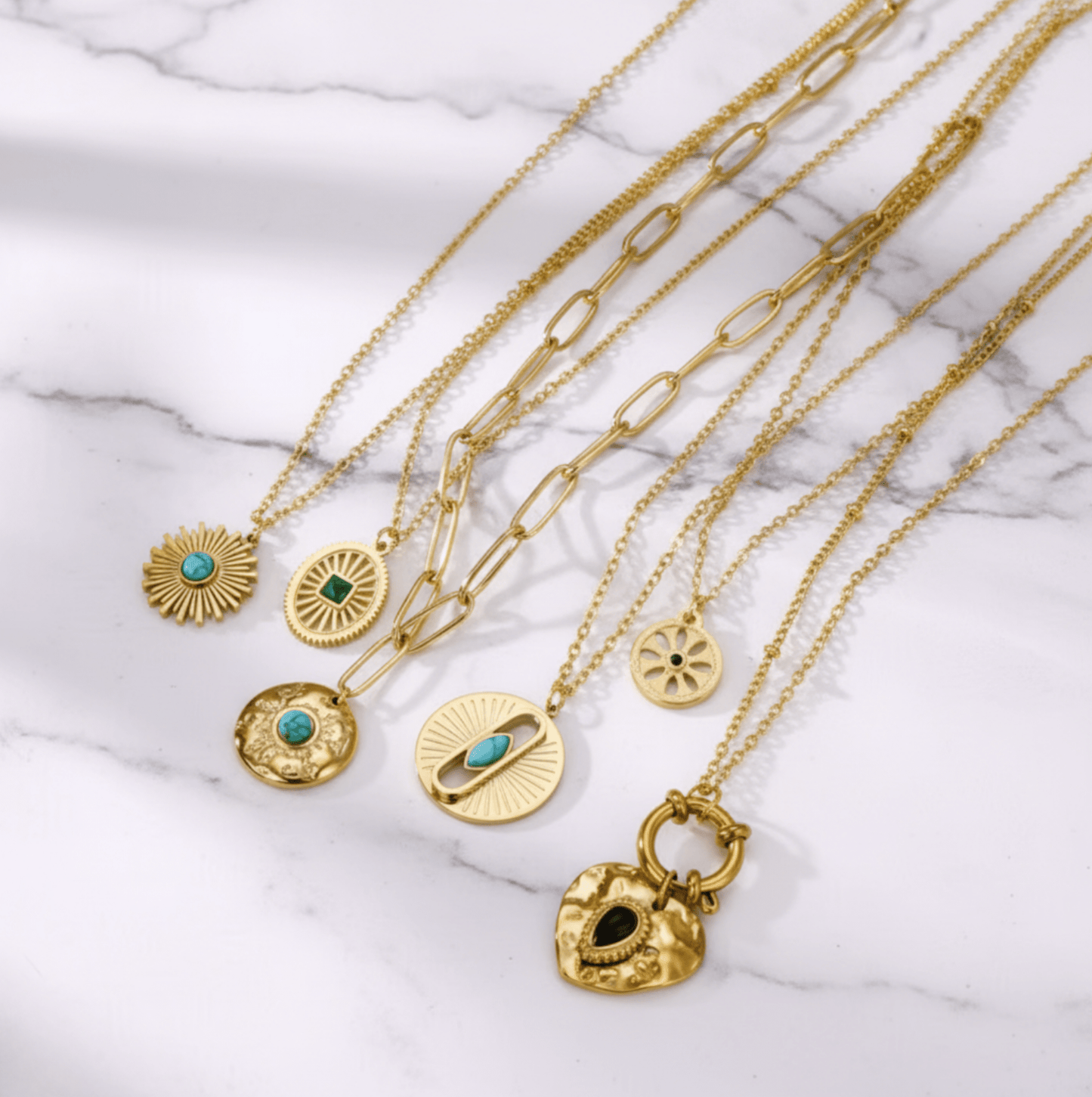 Monerose Soleil Ketting collectie met alle zes varianten op wit marmer, van links Turquoise Zon, Smaragd Ovaal, Paperclip Schakel, Turquoise Oog, Gouden Munt en Zwart Hart, 18K goldplated 316L roestvrij staal, Lumière collectie van Monerose