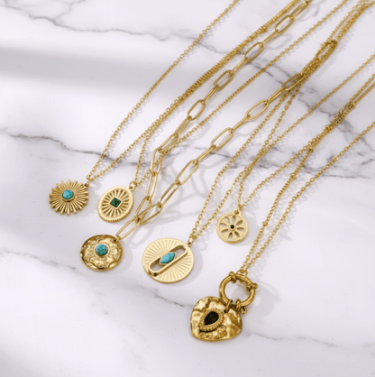 Monerose Soleil Ketting collectie met alle zes varianten op wit marmer, van links Turquoise Zon, Smaragd Ovaal, Paperclip Schakel, Turquoise Oog, Gouden Munt en Zwart Hart, 18K goldplated 316L roestvrij staal, Lumière collectie van Monerose