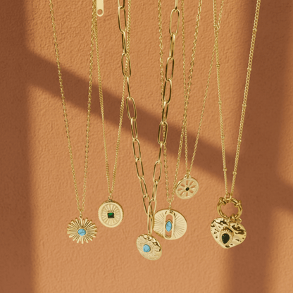 Alle zes varianten van de Monerose Soleil Ketting hangend op warme terracotta achtergrond, 18K goldplated roestvrij staal met turquoise, smaragd en zwarte natuurstenen, Lumière collectie van Monerose