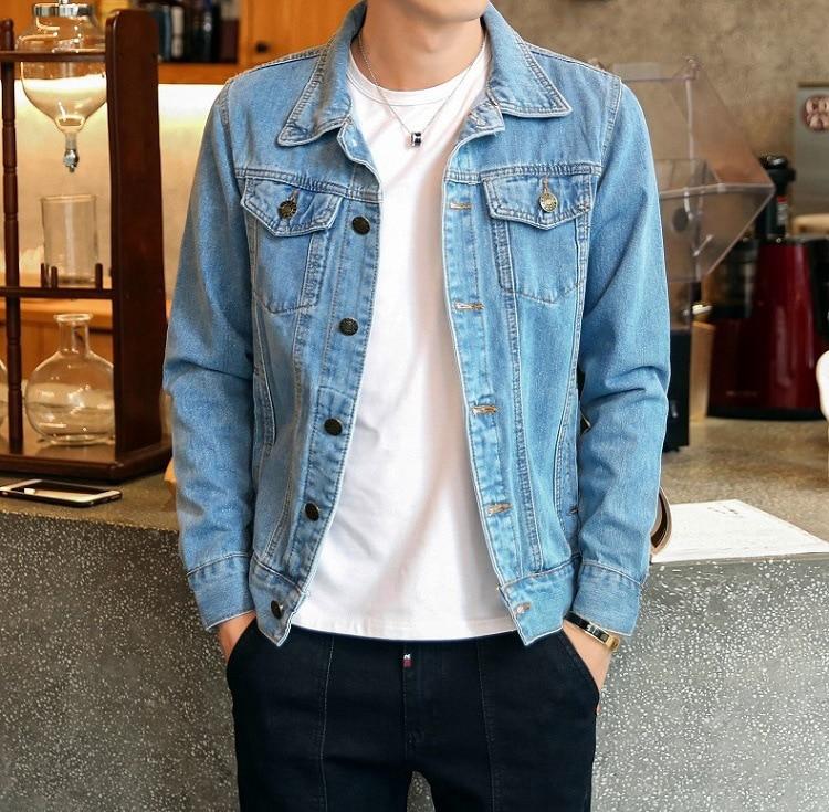 Homme portant une veste en jean bleu clair, chemise blanche et pantalon noir, debout dans un café moderne. Mode décontractée, style urbain.