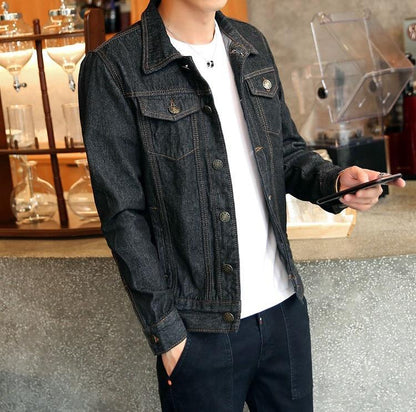 Homme portant une veste en jean noir, debout dans un café moderne, tenant un smartphone. Mode décontractée, style urbain, tendance masculine.