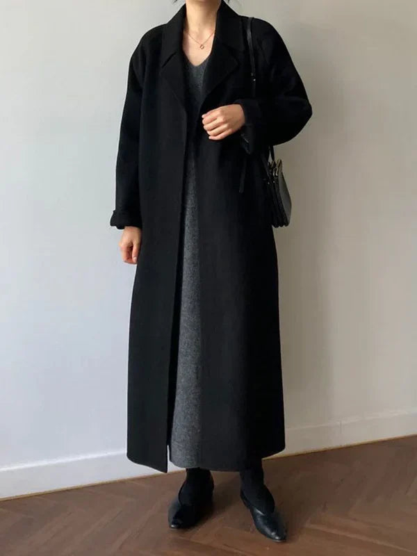 Femme portant un long manteau noir élégant, robe grise, chaussures noires, sac à main noir, posant sur un sol en bois. Mode automne-hiver chic.