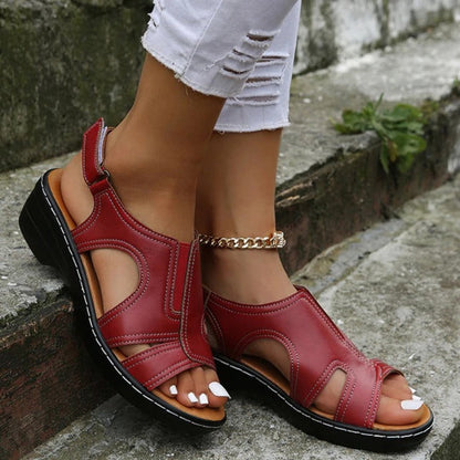 Sandales rouges en cuir pour femme, style décontracté, avec talon bas, portées avec un jean blanc déchiré, sur fond de pierre grise.